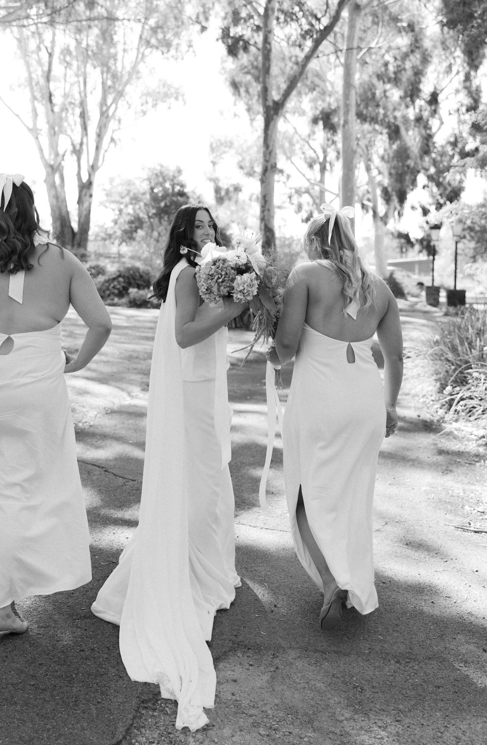 Elegant bridesmaids in white dresses walk down a garden path holding bouquets at an outdoor wedding ceremony.