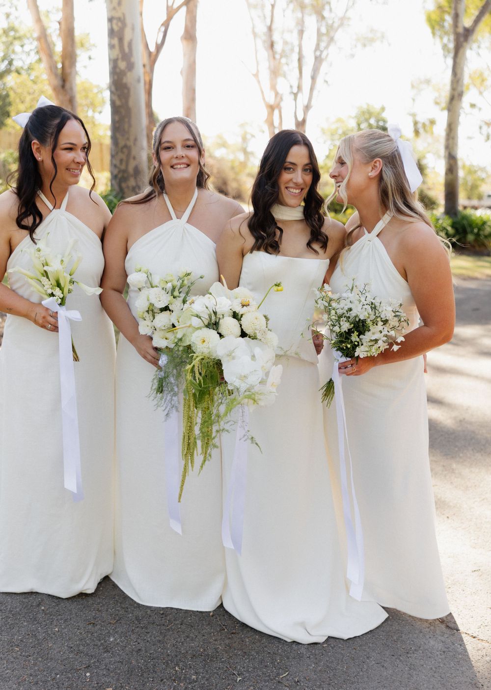 Bridesmaids wearing elegant white halter dresses hold white and green bouquets while smiling together outdoors.