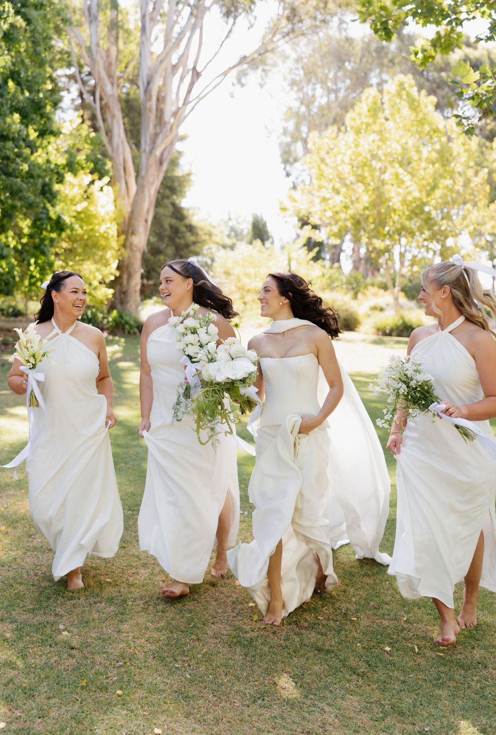 Bridesmaids in flowing white dresses run joyfully through a sunlit garden while holding bouquets.