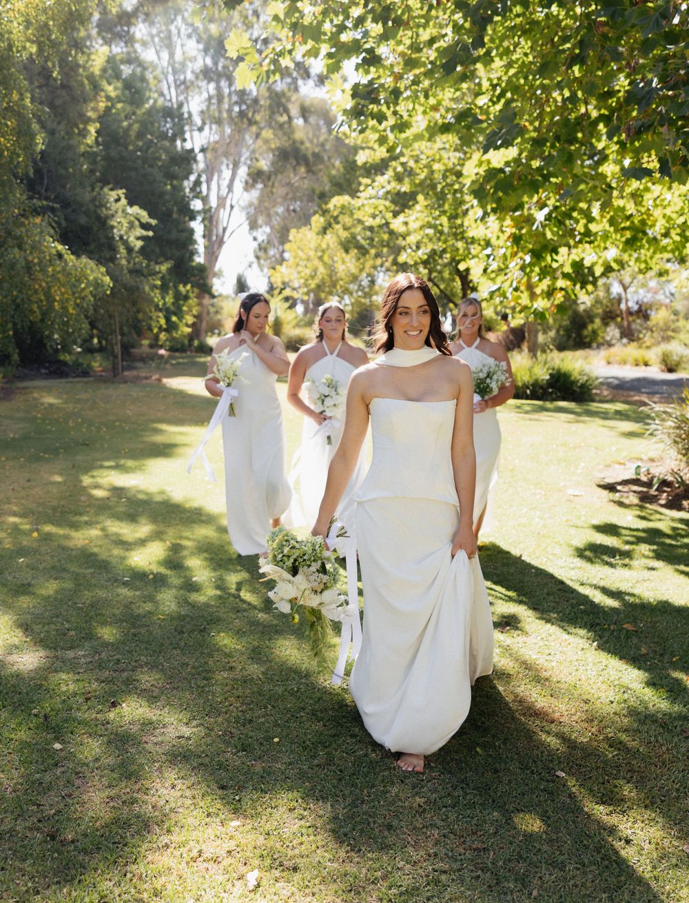 A group in flowing white dresses walks along a sunlit garden path lined with lush greenery and trees.