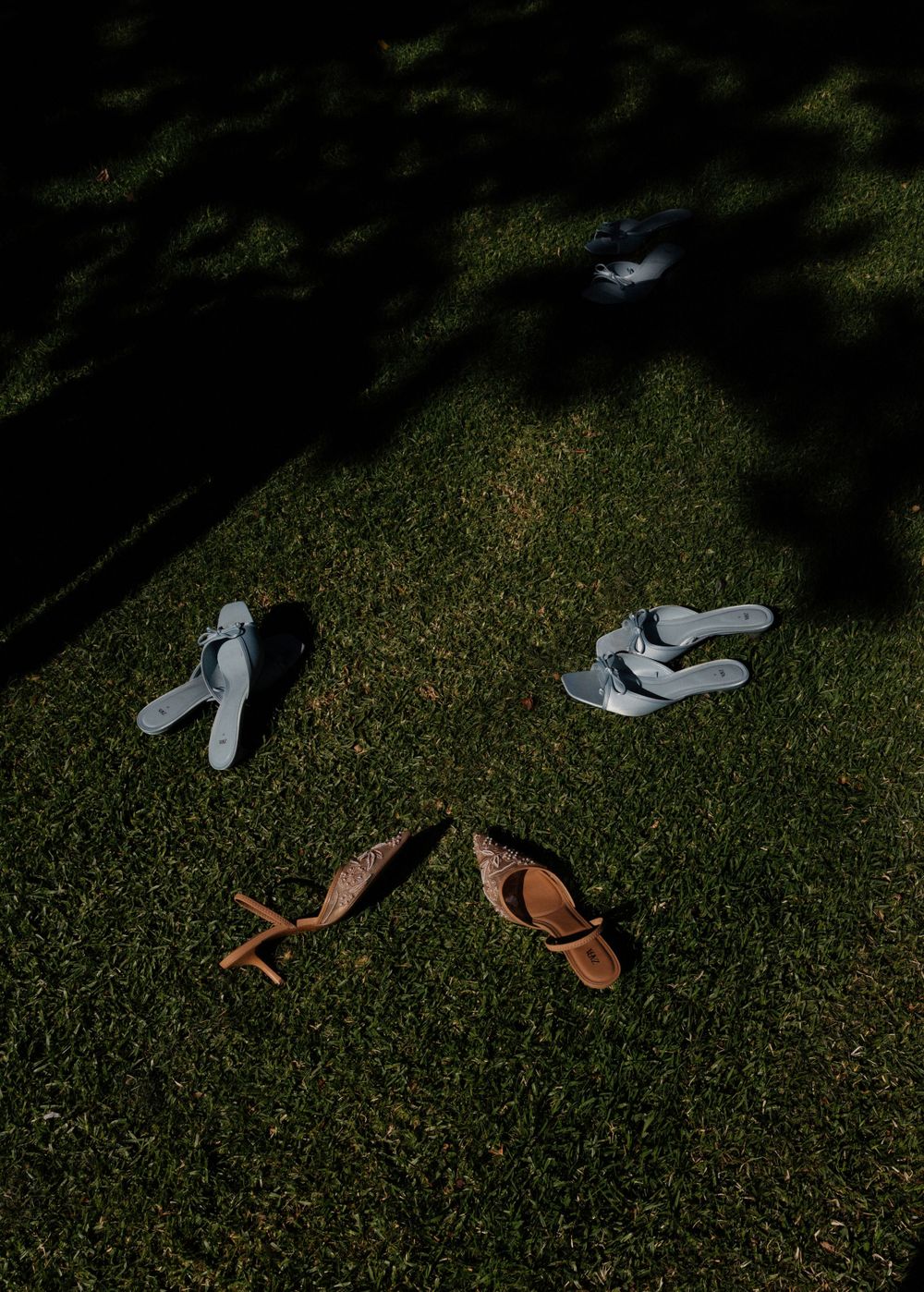 Wedding shoes and sandals arranged on grass in shadow and sunlight.