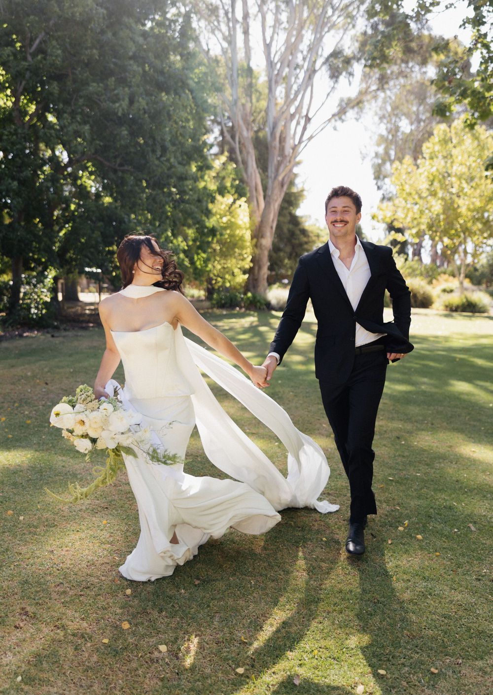 A newlywed couple runs joyfully through a lush garden as the bride's dress flows in the wind.