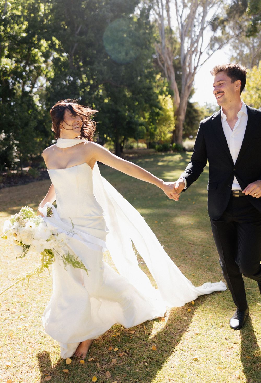 A joyful couple in wedding attire dance and laugh together on a sunlit lawn surrounded by trees.