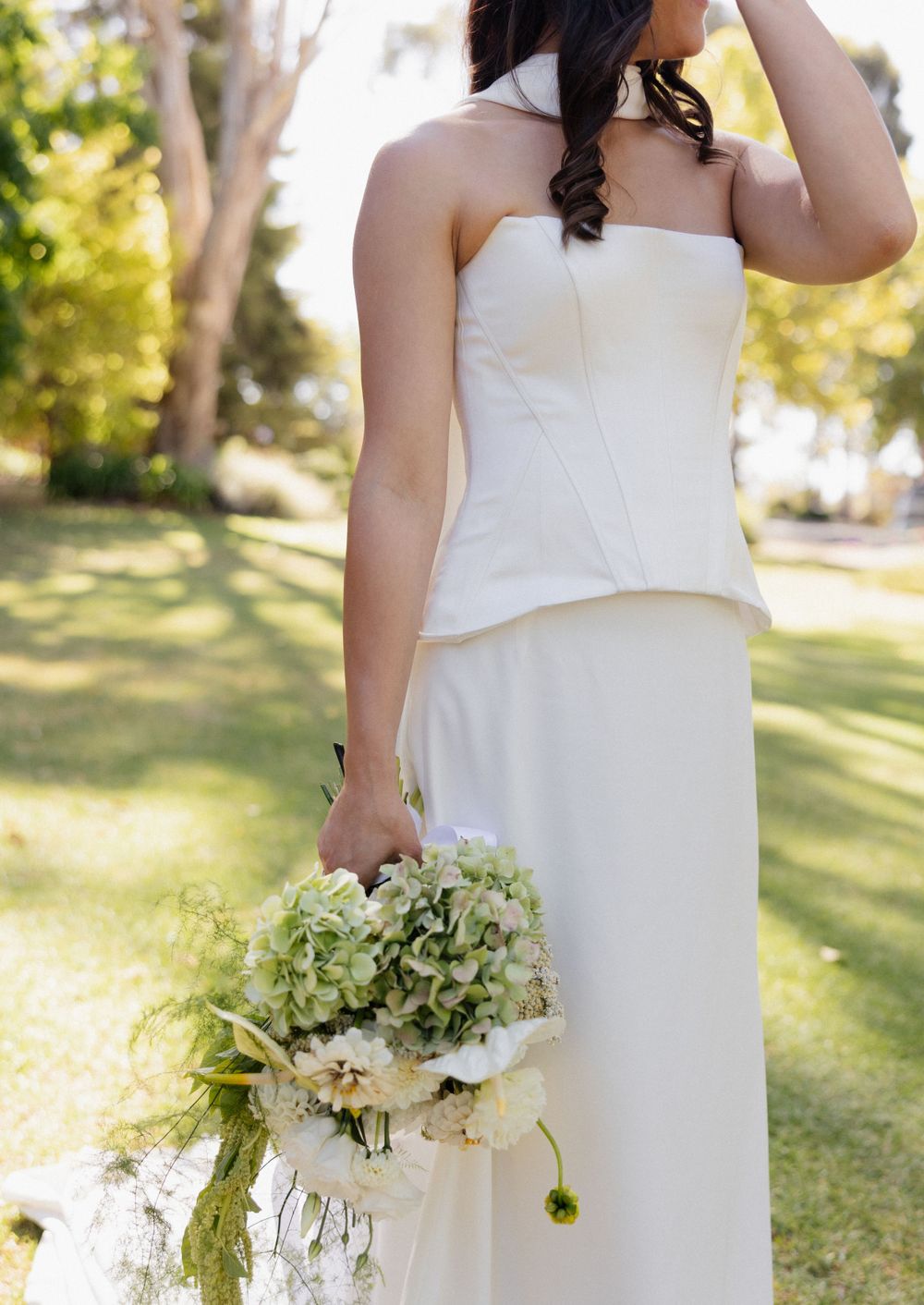 An elegant white wedding dress and bouquet of hydrangeas photographed in a sunny outdoor garden setting.