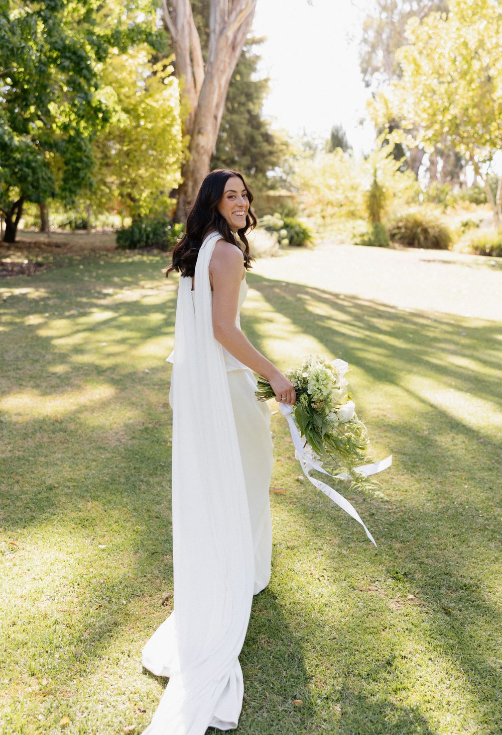 A person in a flowing white wedding dress holds a delicate bouquet of flowers in a sun-dappled garden setting.