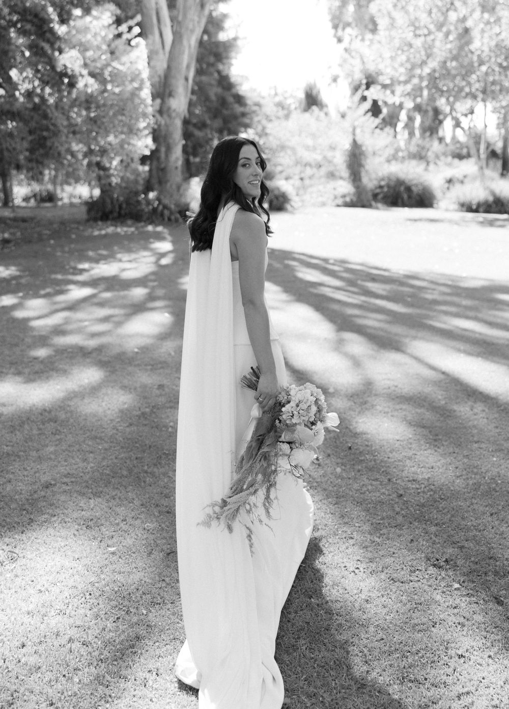 A black and white portrait of a bride in a sleek modern wedding gown holding a bouquet in a garden.