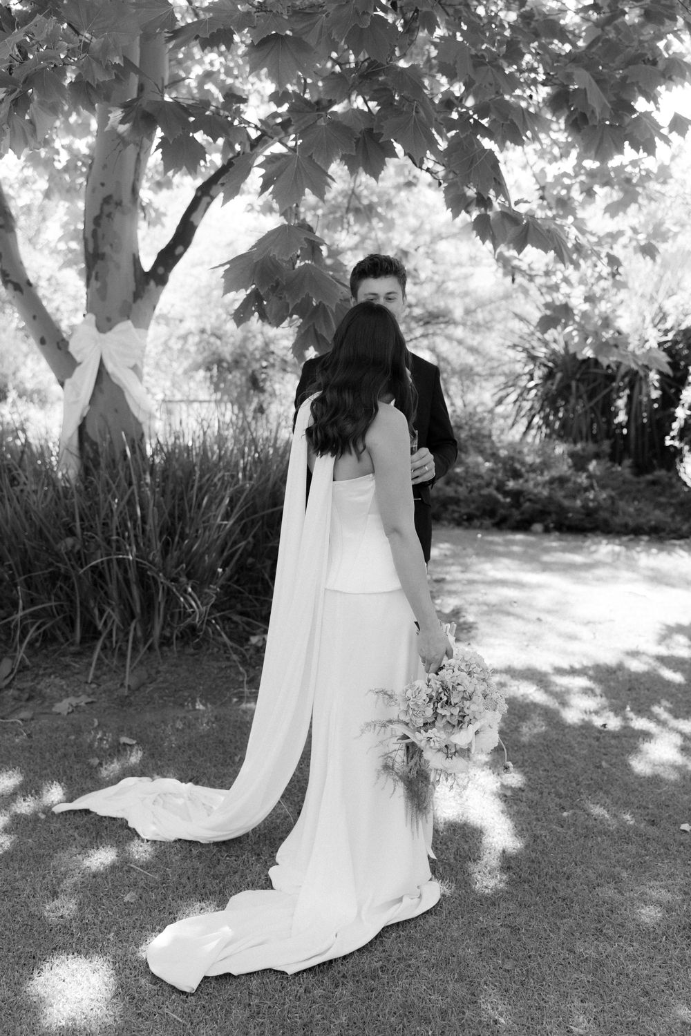 Black and white photo of a wedding couple embracing beneath a leafy tree in a garden setting.