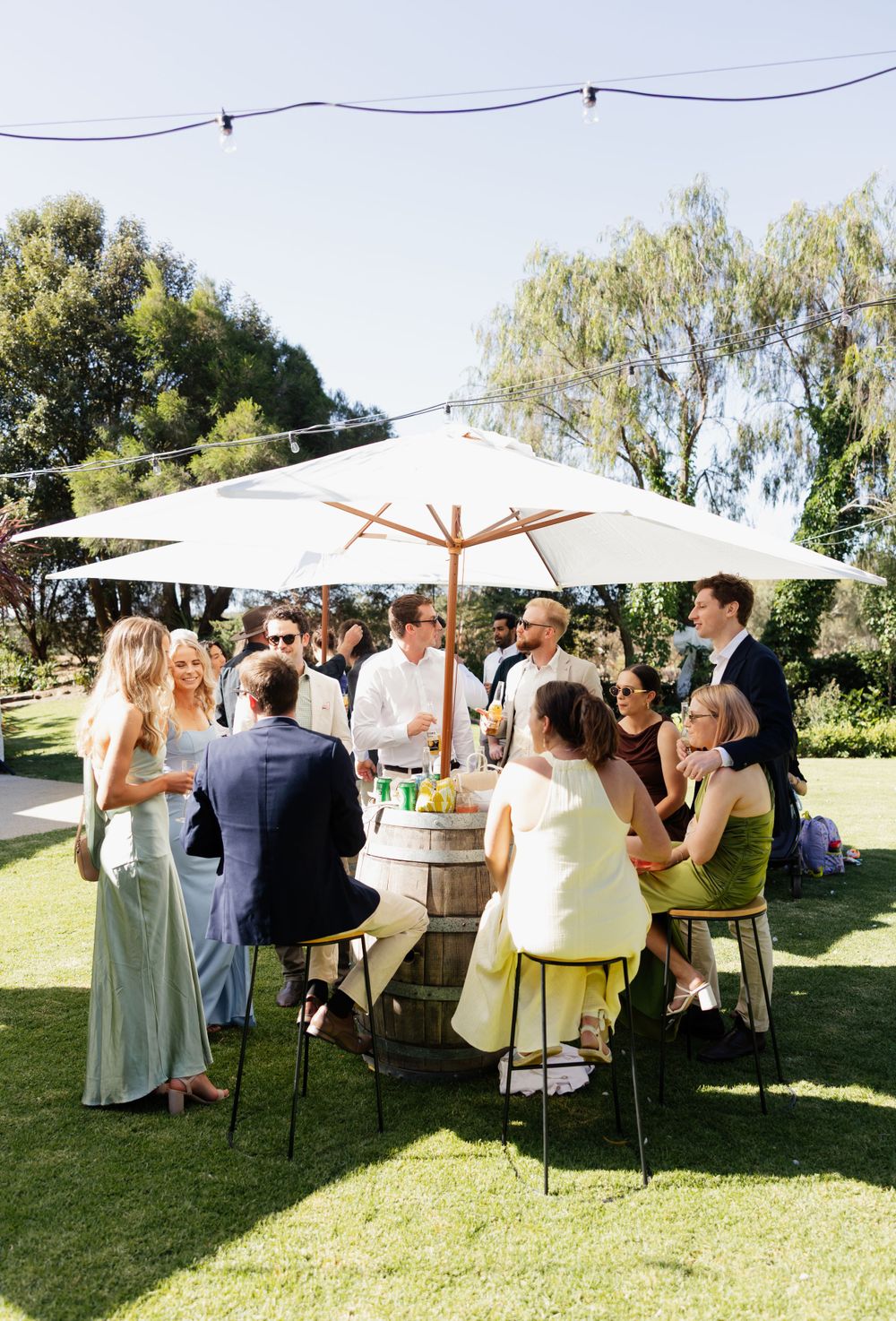 Group of guests gather around wine barrels under white market umbrellas at an outdoor garden party on a sunny day.