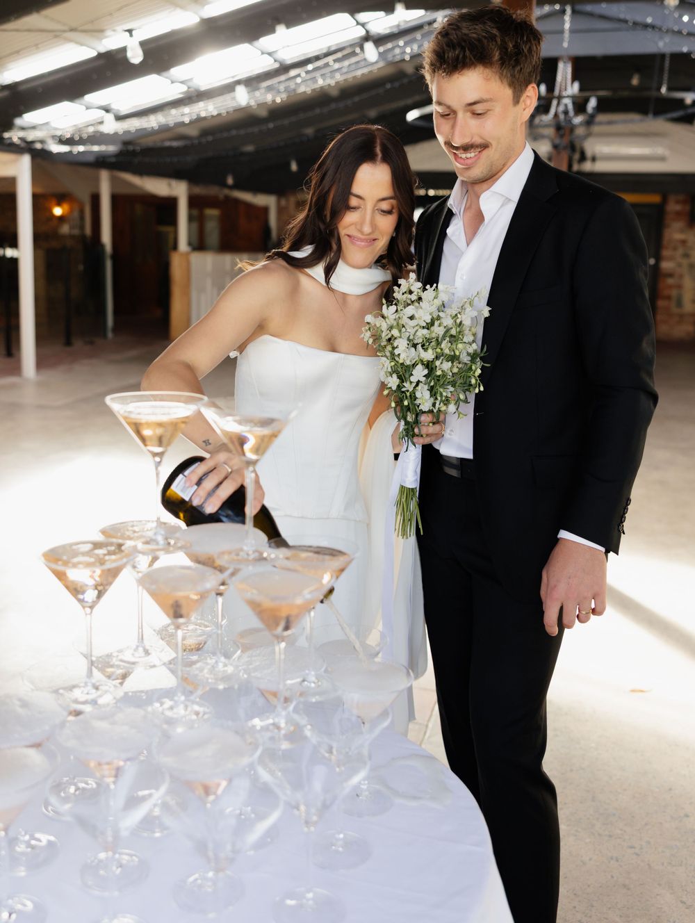 A couple at their wedding reception pour champagne into a tower of elegant martini glasses holding a white bouquet.