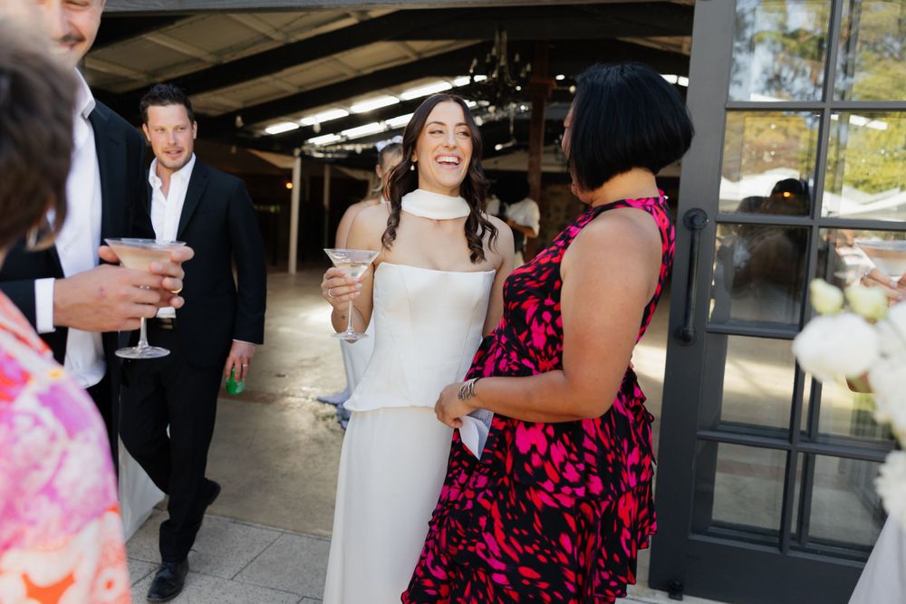A bride in a white dress laughs joyfully while socializing with guests at an outdoor reception.
