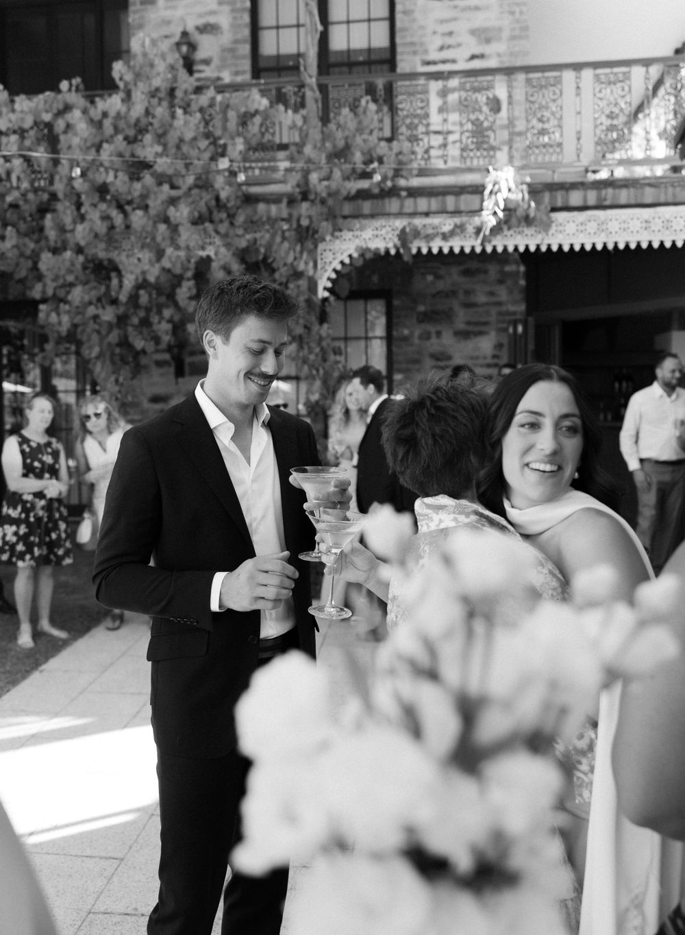 Black and white photograph showing an elegant event with blossoming trees and formal attire in an outdoor venue.