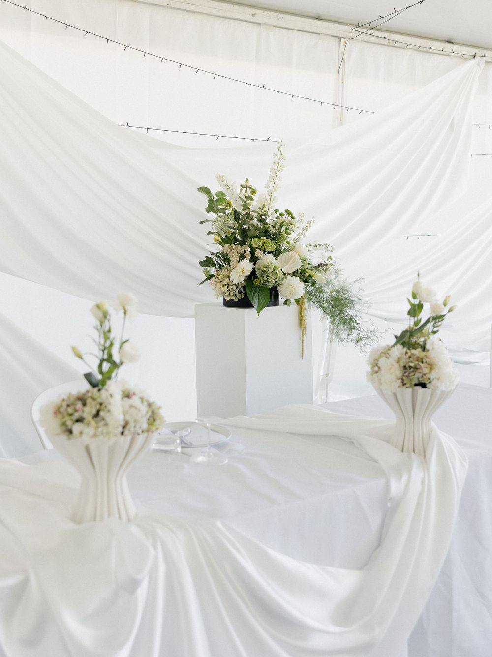 Elegant white wedding decor setup with floral arrangements in white vases against a draped white backdrop.