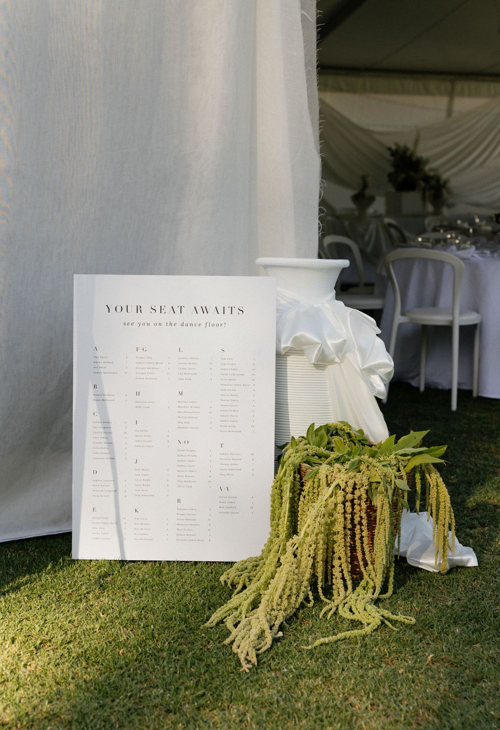 Wedding seating chart display with decorative greenery and white draping in an outdoor tent reception setting.