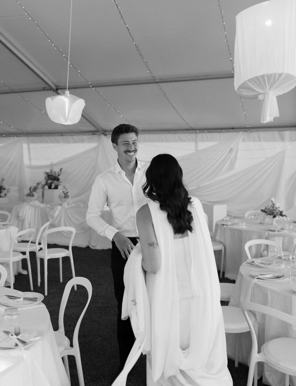 Two people share a moment together in a white tent reception venue with elegant tables and hanging lantern decorations.
