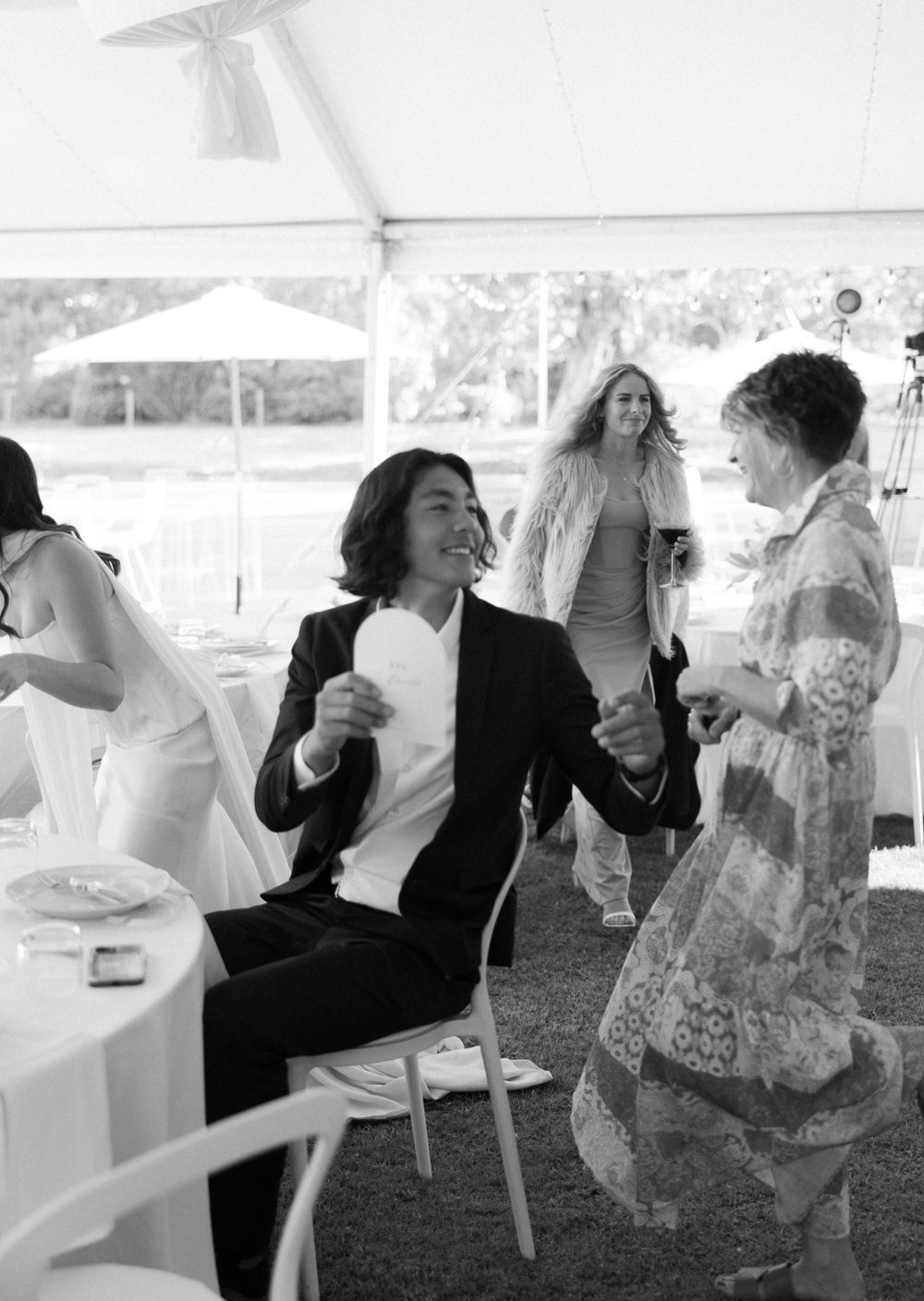A candid black and white photo of guests enjoying conversation and laughter under a white tent at an outdoor event.
