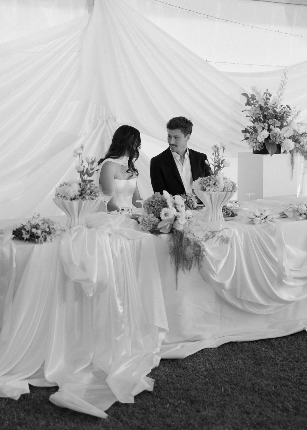 Black and white photo of a wedding couple seated at an elegantly draped table with floral arrangements under a white tent.