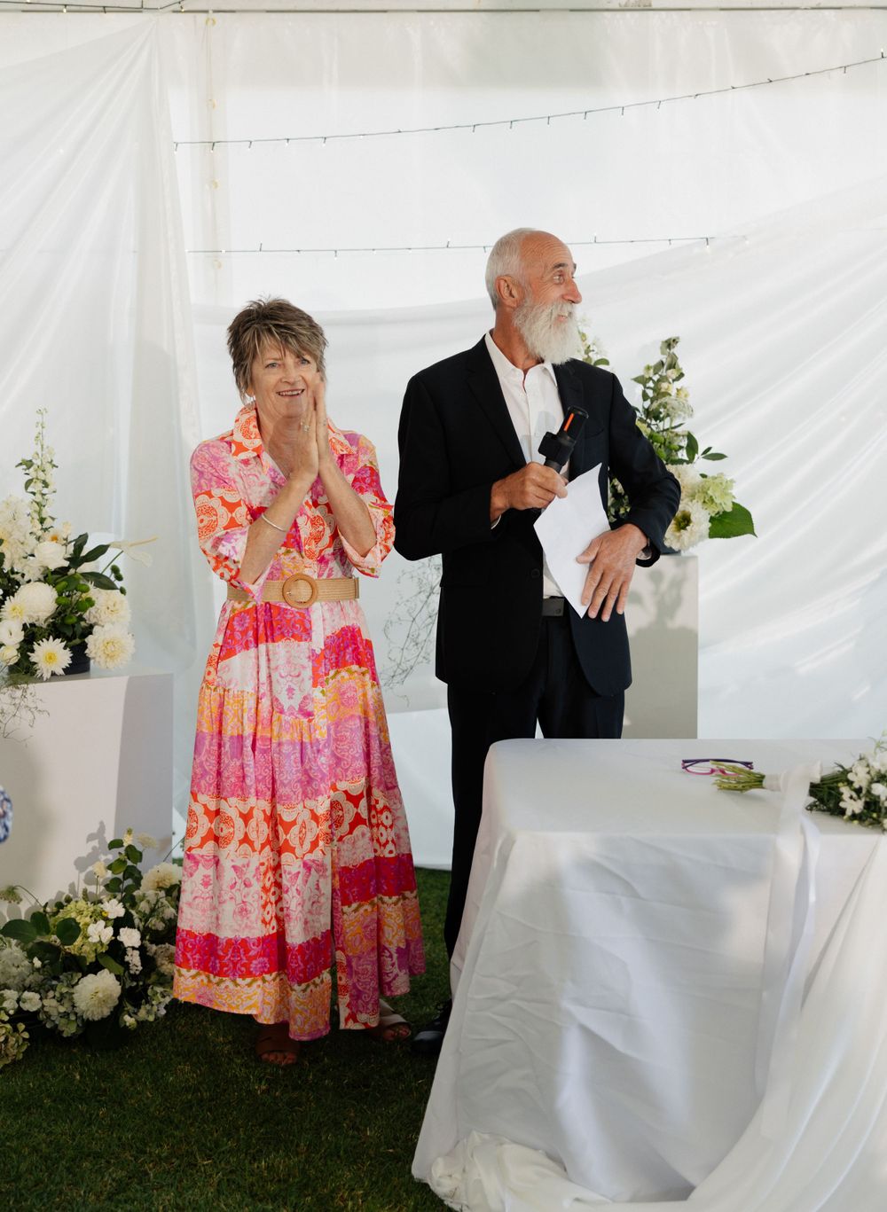 Two people stand in front of white drapes at a wedding reception, with one wearing a colorful pink dress and one in a tuxedo.