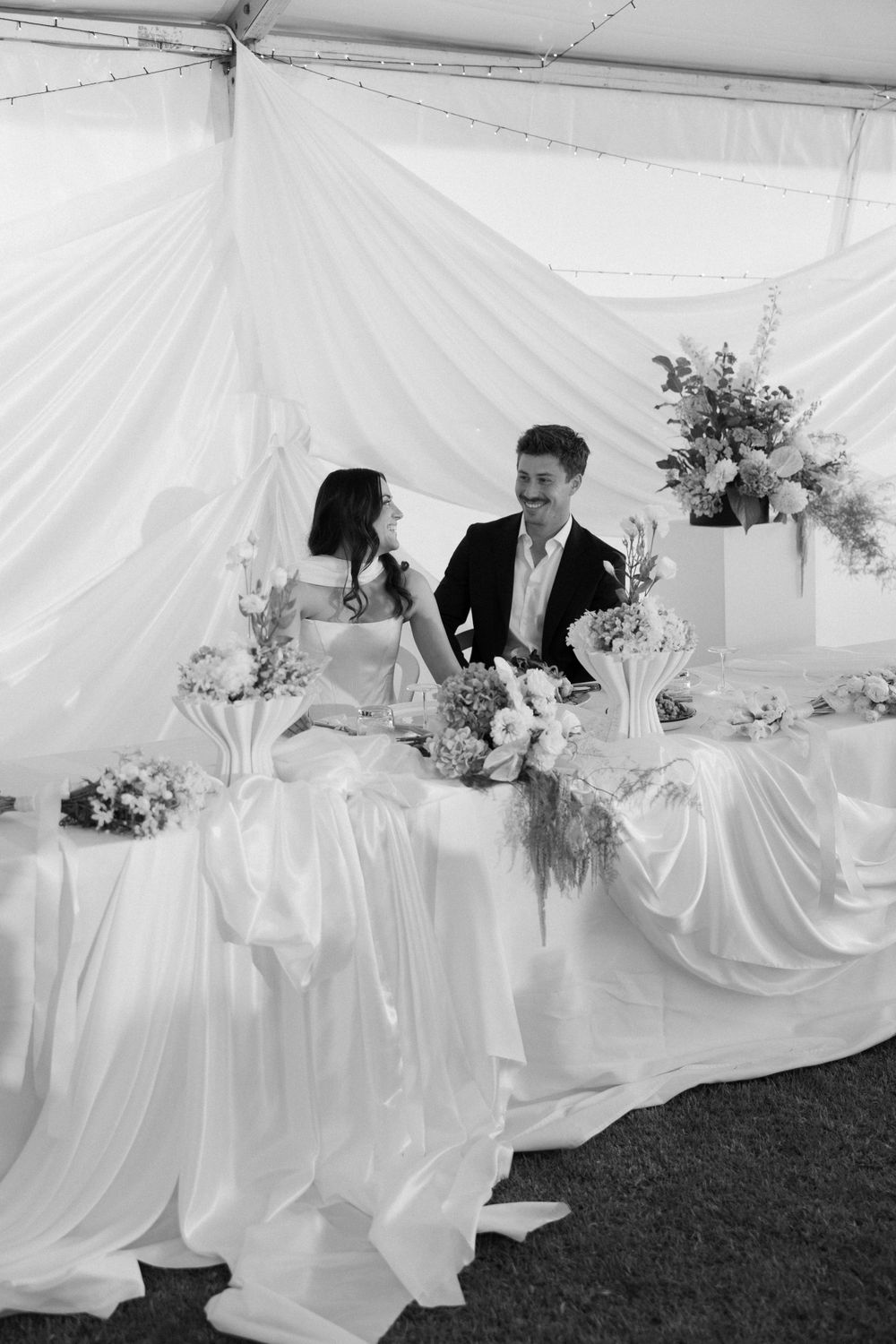 A newlywed couple sits at an elegant white draped wedding reception table under a tent with floral arrangements.
