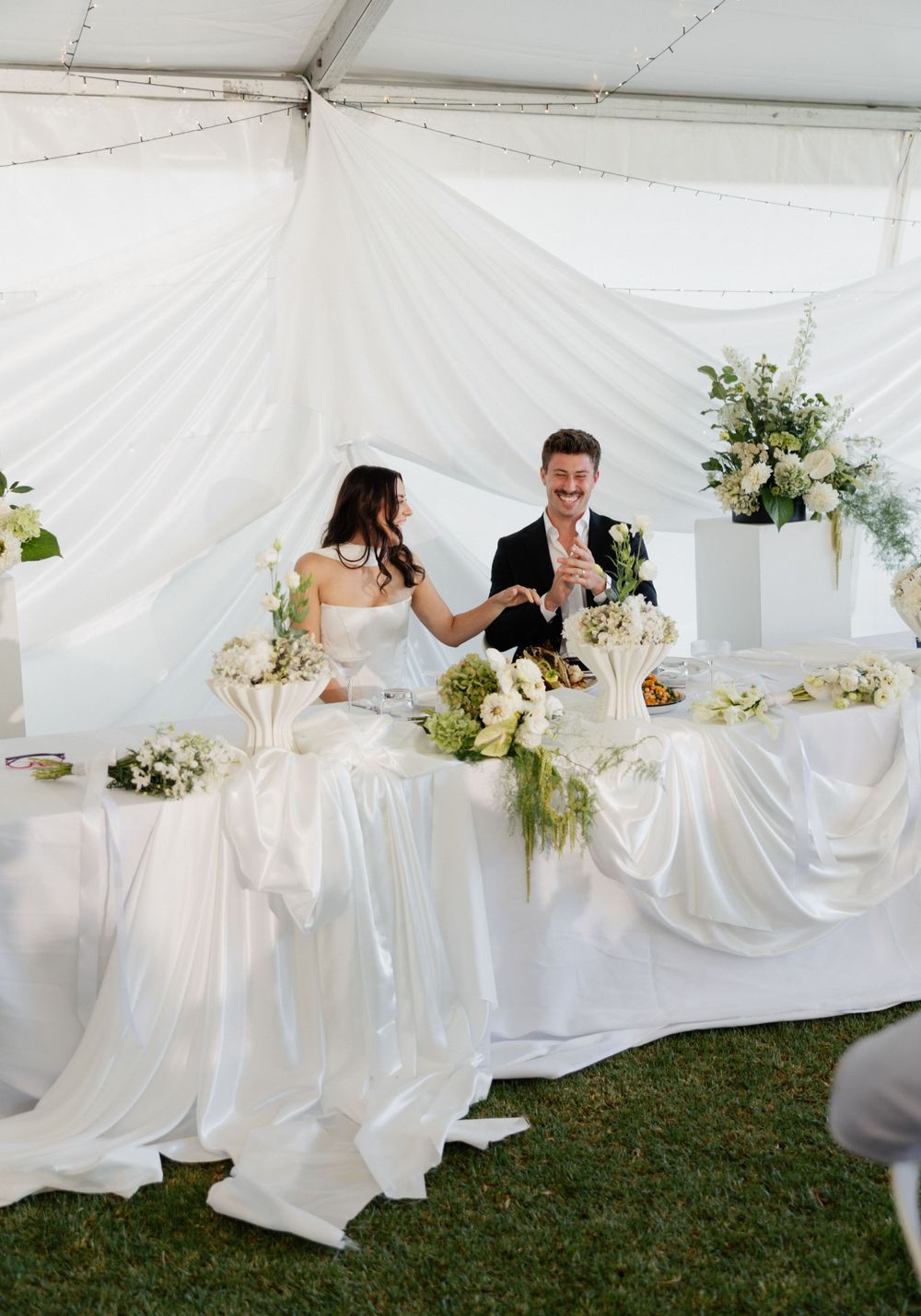 A couple sits at an elegant white-draped wedding table under a tent with floral arrangements and satin tablecloths.