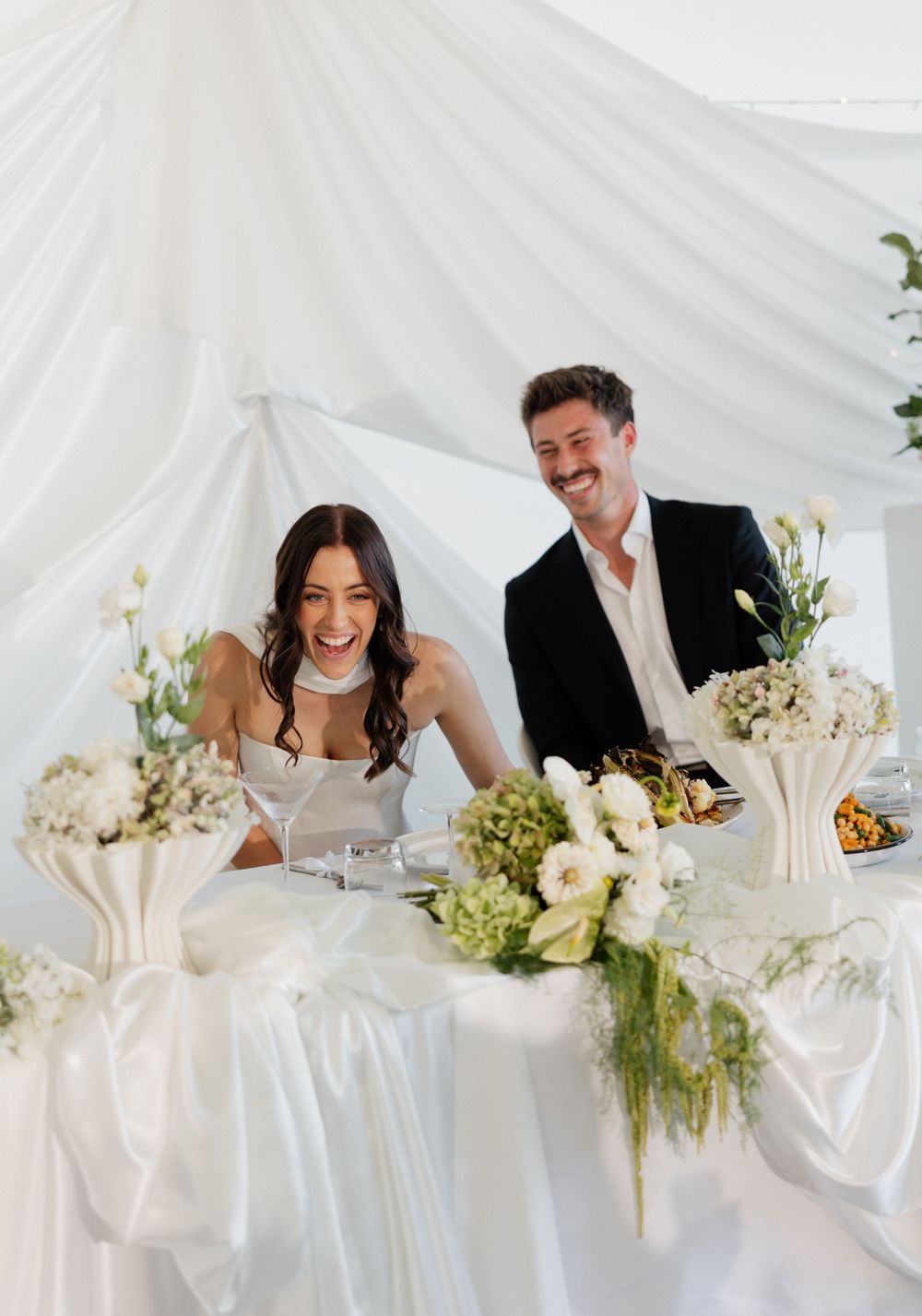 A joyous wedding reception moment with newlyweds laughing together at a white-decorated table with floral arrangements.
