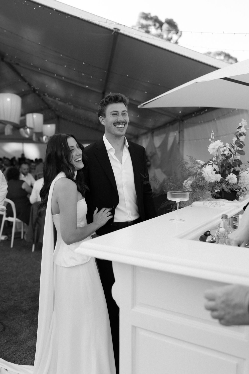 Black and white photo of newlyweds sharing a laugh at their outdoor reception by a white bar counter.