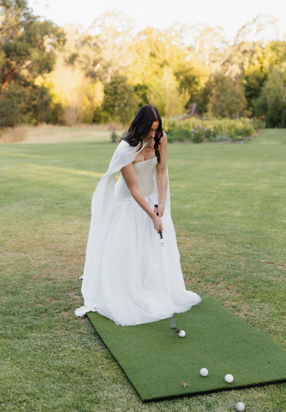 A person in a flowing white wedding dress practices putting on a portable golf mat during an outdoor golden hour photo shoot.