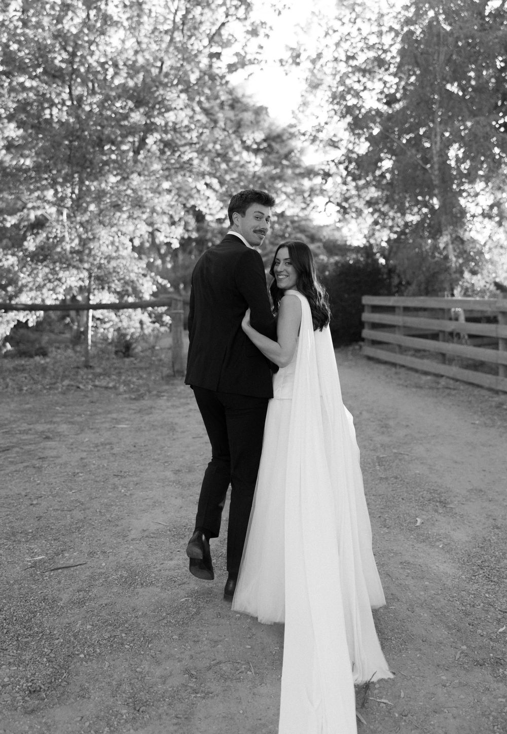 A romantic black and white wedding photo showing a couple embracing on a dirt path surrounded by trees.