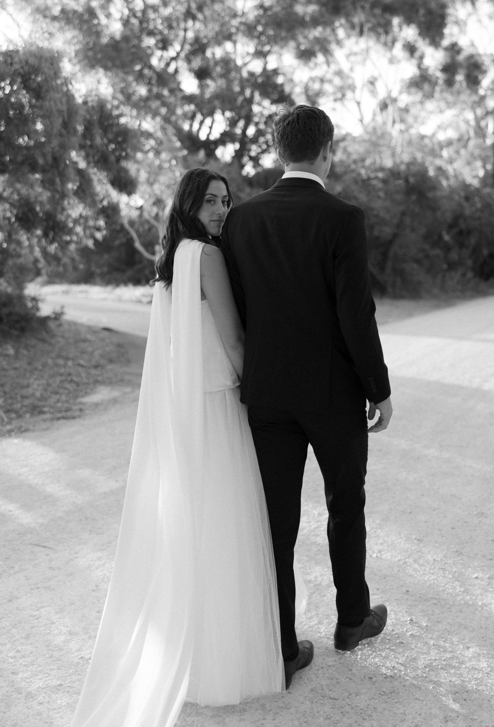 A black and white photo of newlyweds walking together on a path, with the bride's long veil flowing behind them.