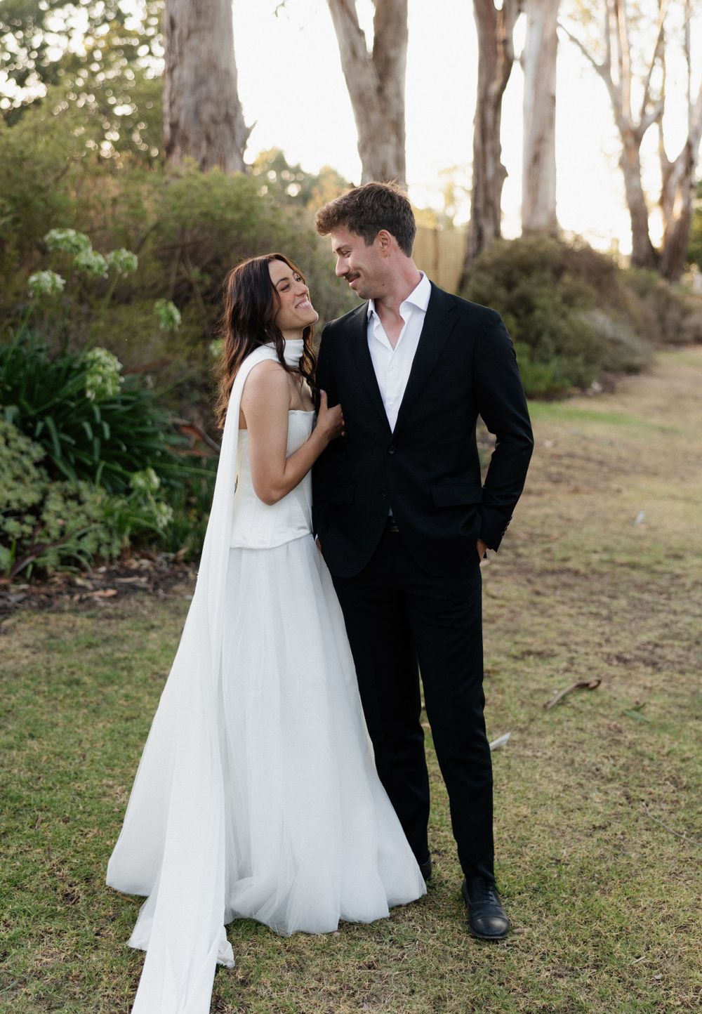 A wedding couple shares a tender moment outdoors among eucalyptus trees, with the bride in white and groom in black.