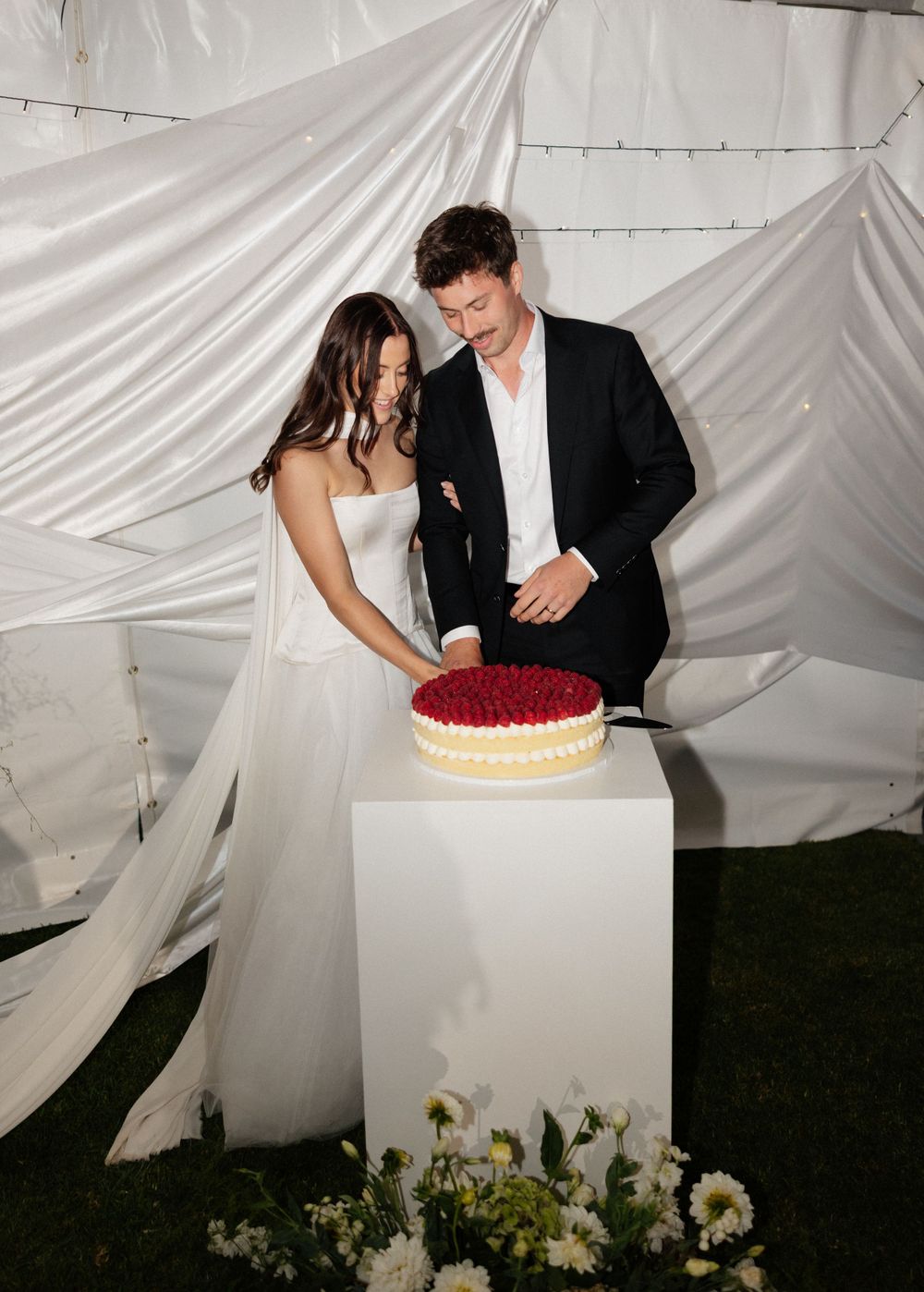 Newlyweds cutting into a red dessert during their wedding reception against a white draped backdrop.