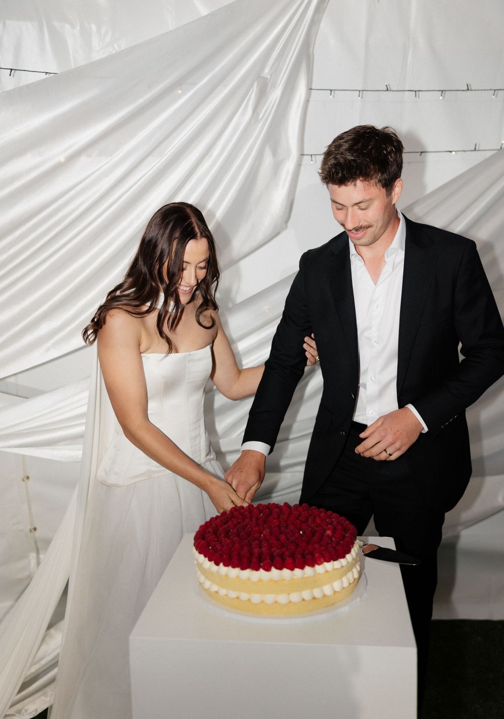 A couple in wedding attire stand together cutting a white cake decorated with red berries in front of a white backdrop.