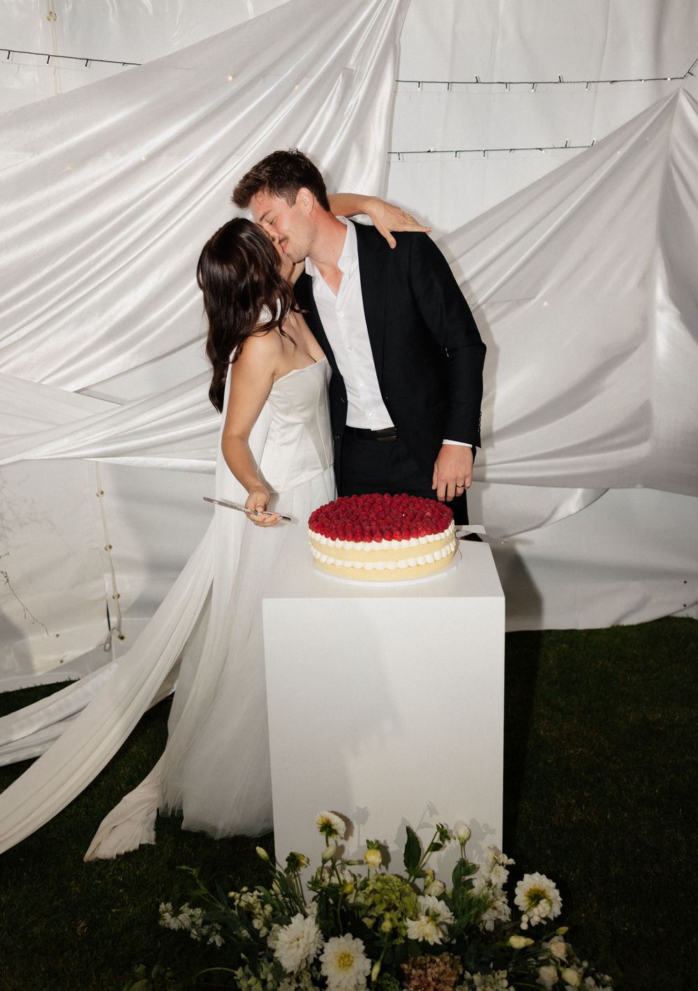 Romantic couple shares intimate moment beside a white pedestal with red-topped cake and white floral decorations.