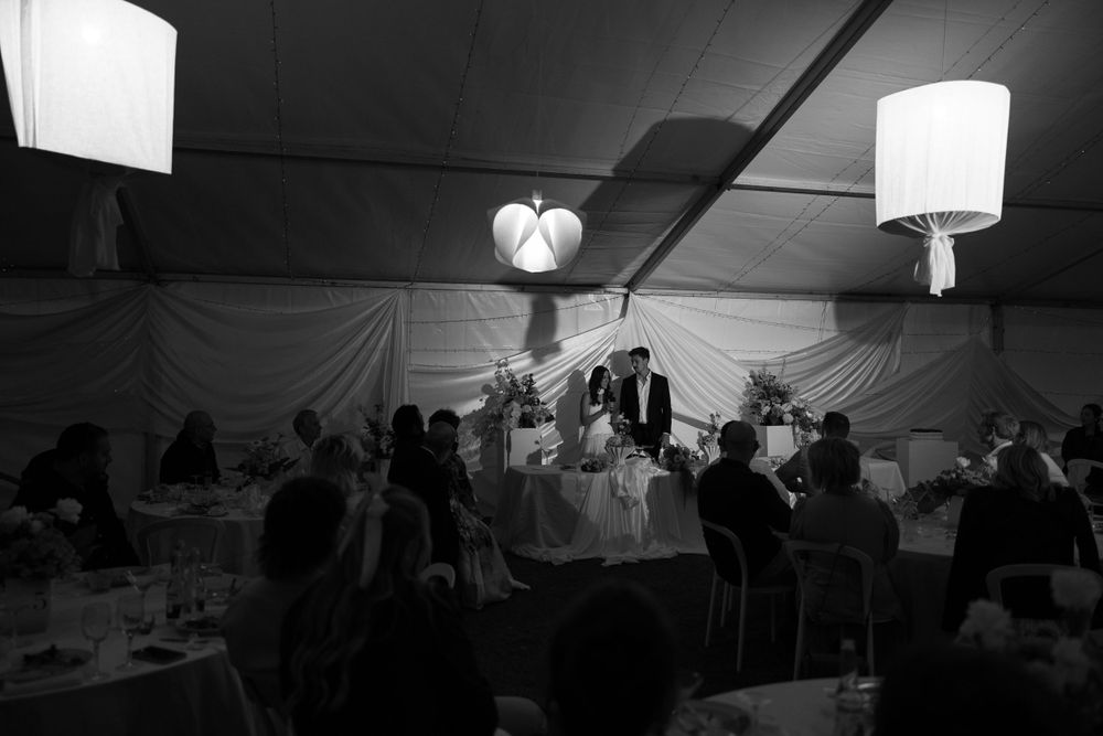 Moody black and white image of a dinner event under a tent with pendant lighting and gathered guests.