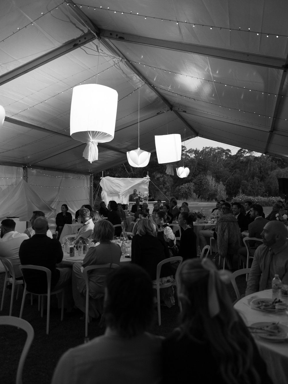 A crowded wedding reception under a white tent with hanging paper lanterns illuminating the dining space.
