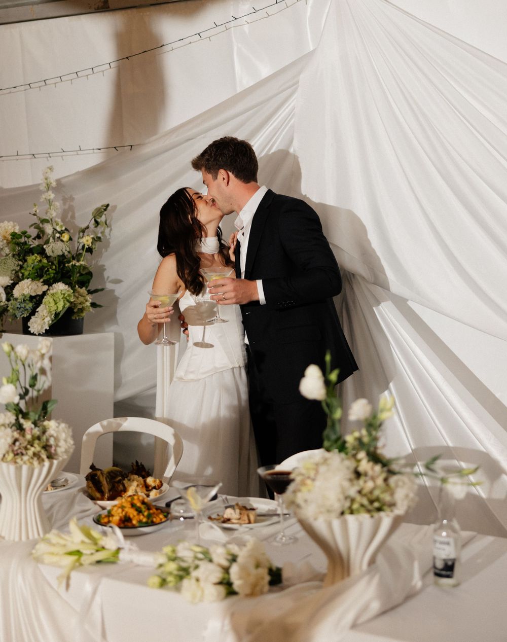 Elegant wedding reception table decorated with white flowers and linens under a draped tent with couple sharing a romantic moment.