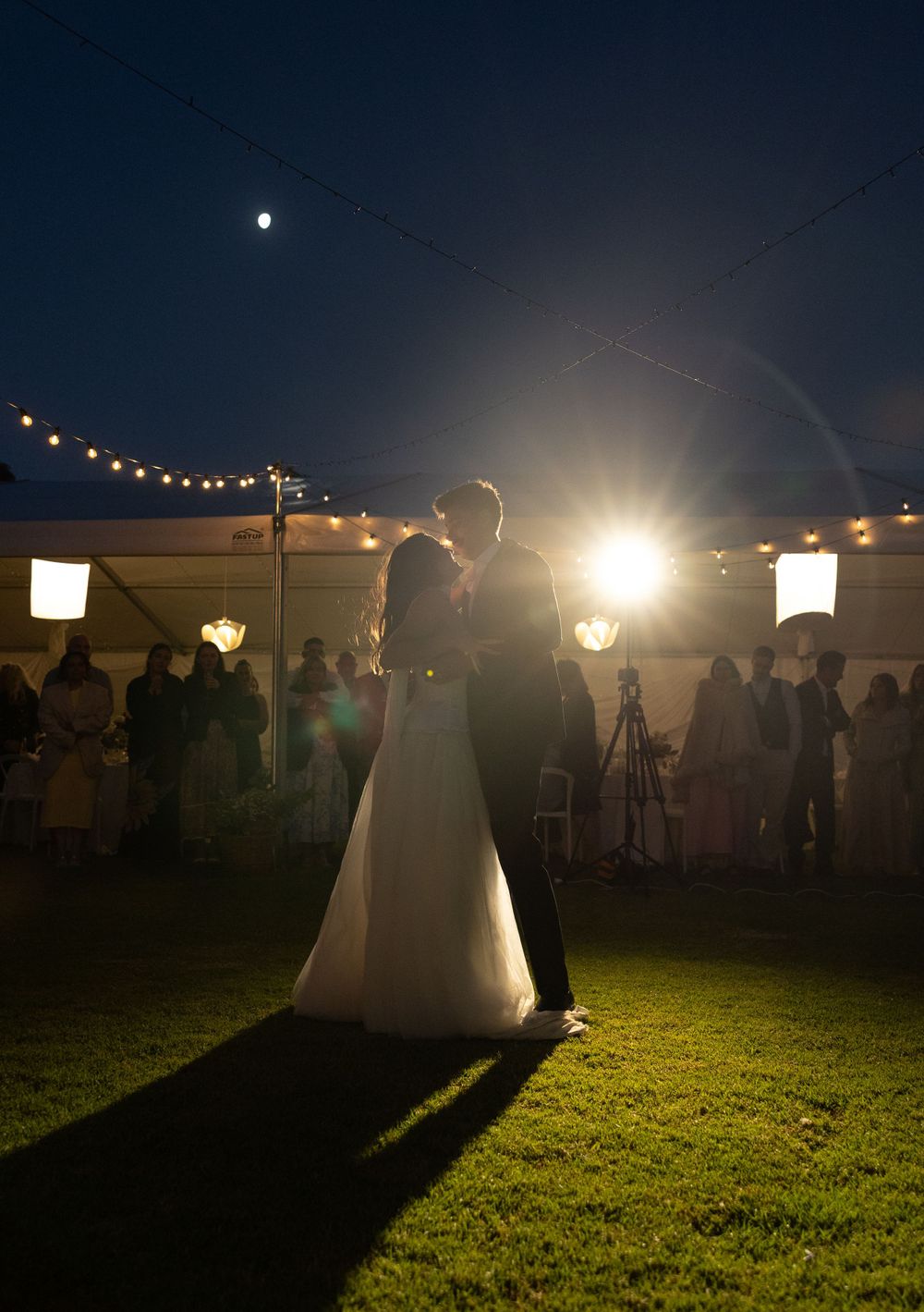 A couple shares their first dance at an outdoor evening wedding reception under string lights and lanterns.