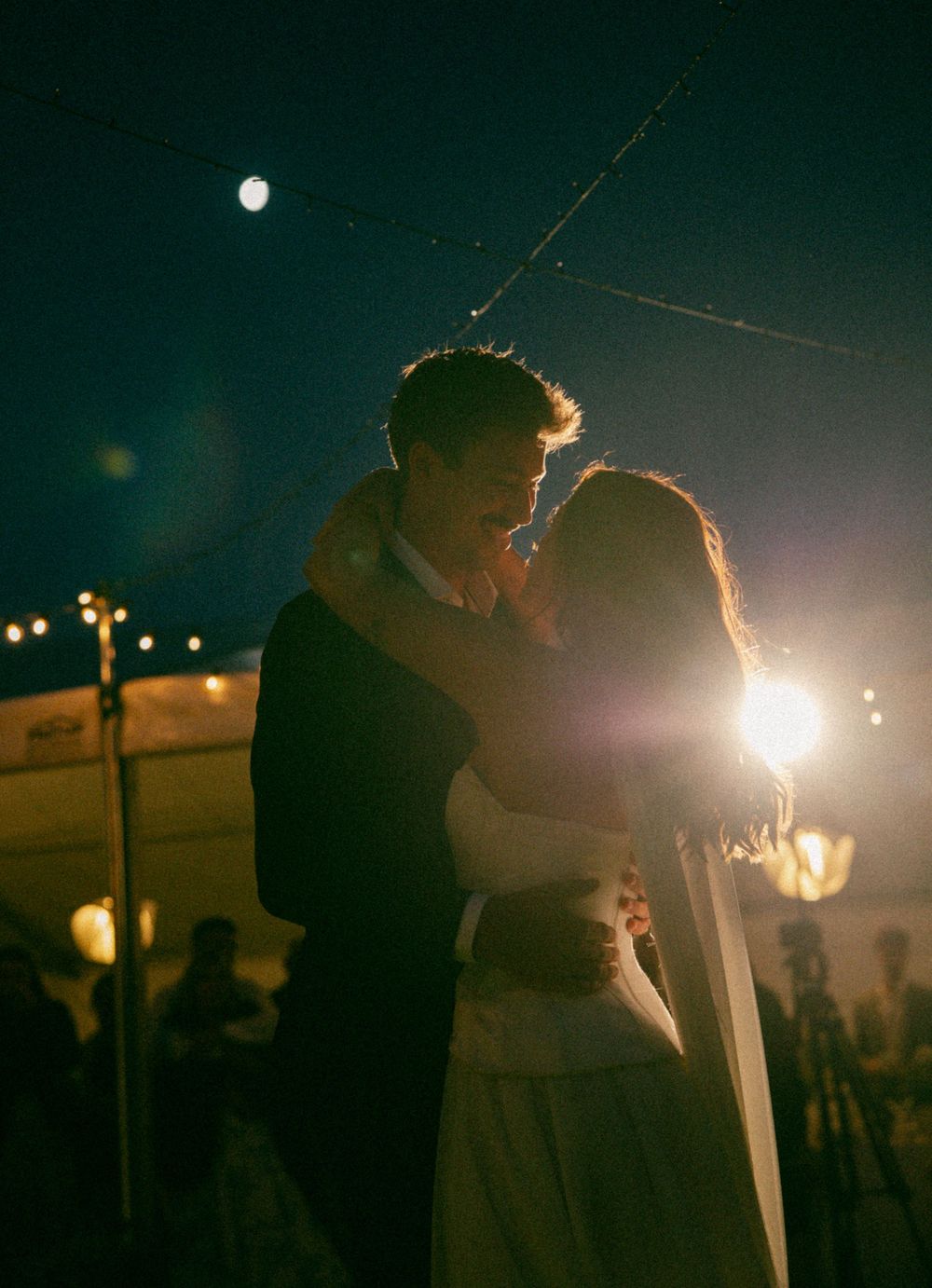 A romantic nighttime embrace under twinkling lights and a full moon at an outdoor wedding celebration.