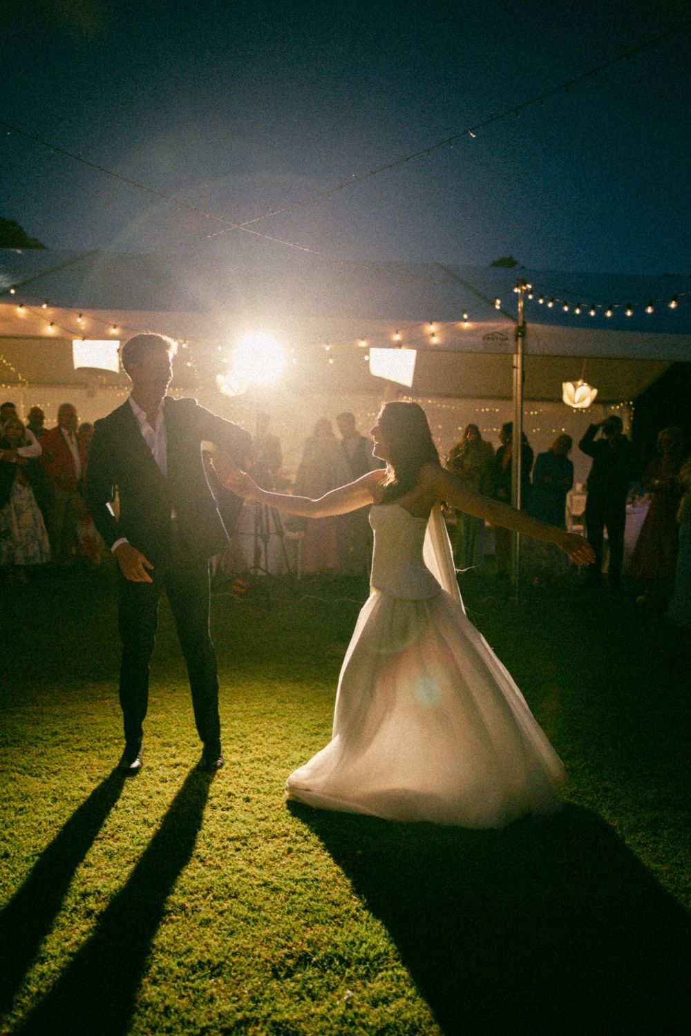 Newlyweds dancing under bright lights at a nighttime outdoor reception with string lights overhead.
