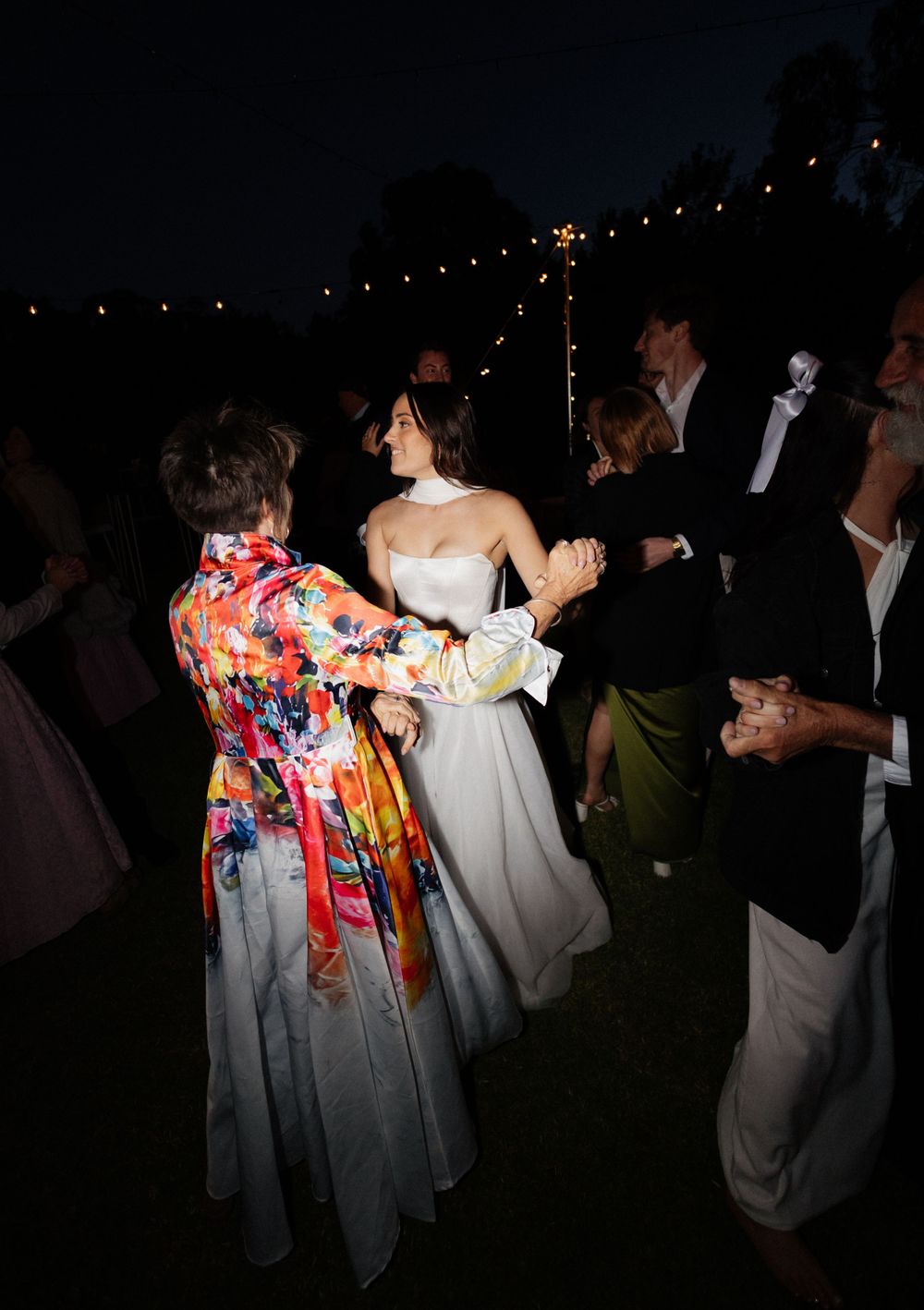 Wedding guests dance together at a nighttime outdoor reception under string lights with a bride in white dress.