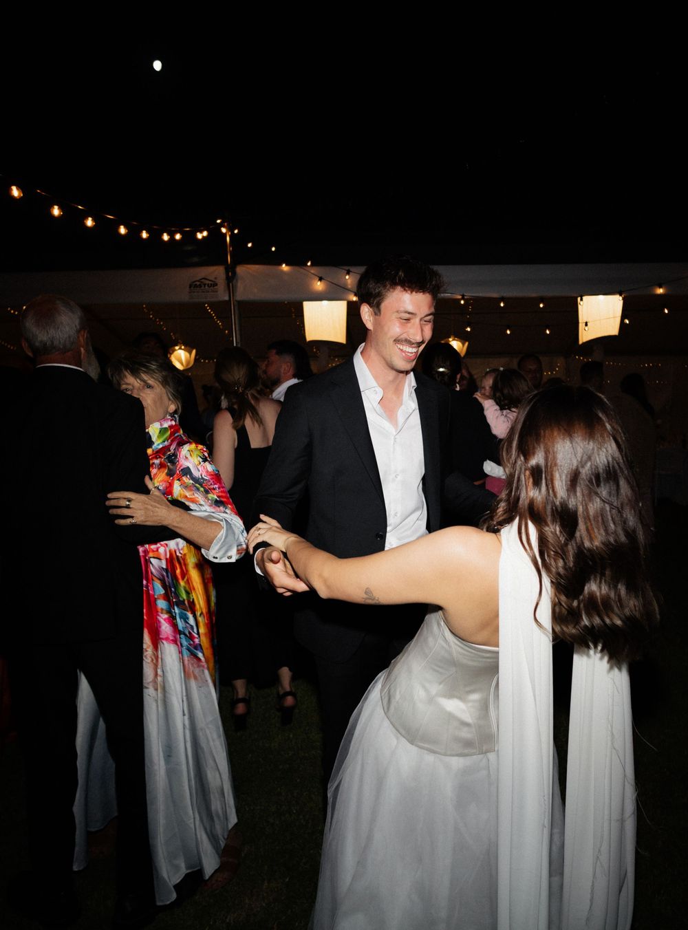 Wedding guests dancing and celebrating during an outdoor evening reception with string lights.