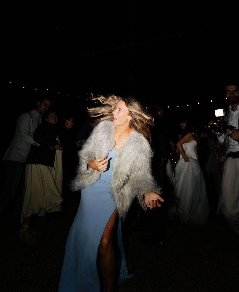 Person in a blue evening gown and furry white jacket dances energetically at a nighttime party with guests in background.