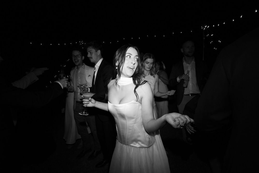 A person in a light dress dances joyfully at a nighttime celebration with string lights glowing in the background.