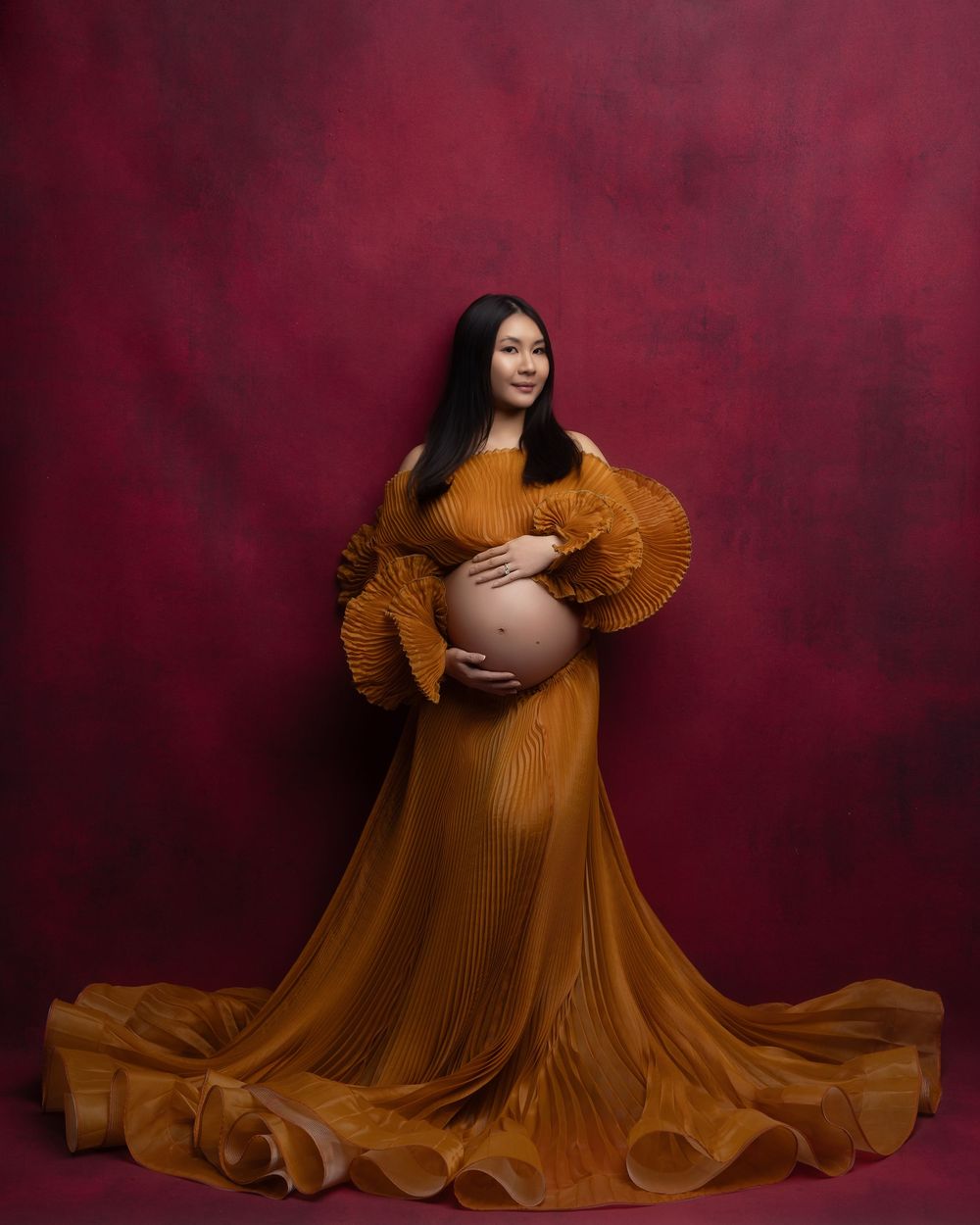 Elegant maternity portrait featuring a flowing mustard yellow gown against a deep burgundy background.