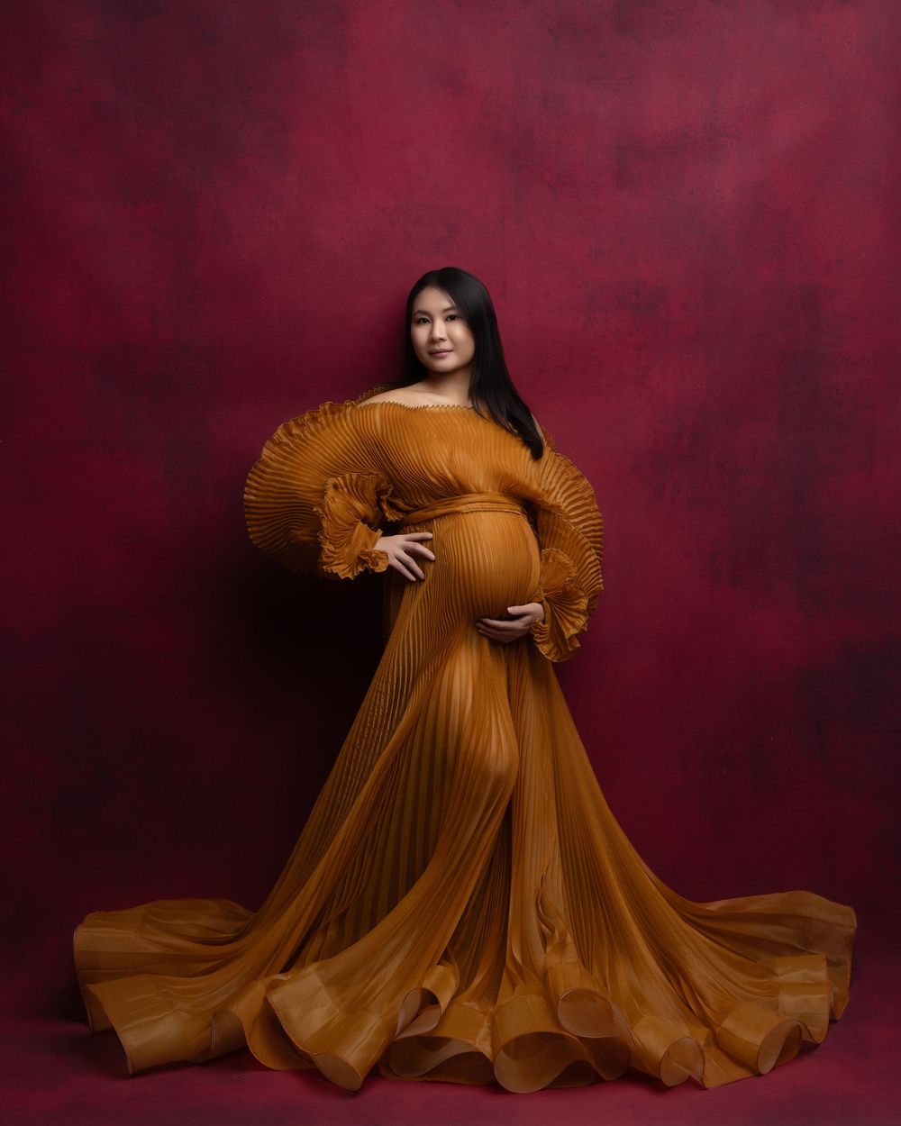 A stunning maternity photo series in a flowing burnt orange gown against a deep burgundy backdrop creates an ethereal mood.