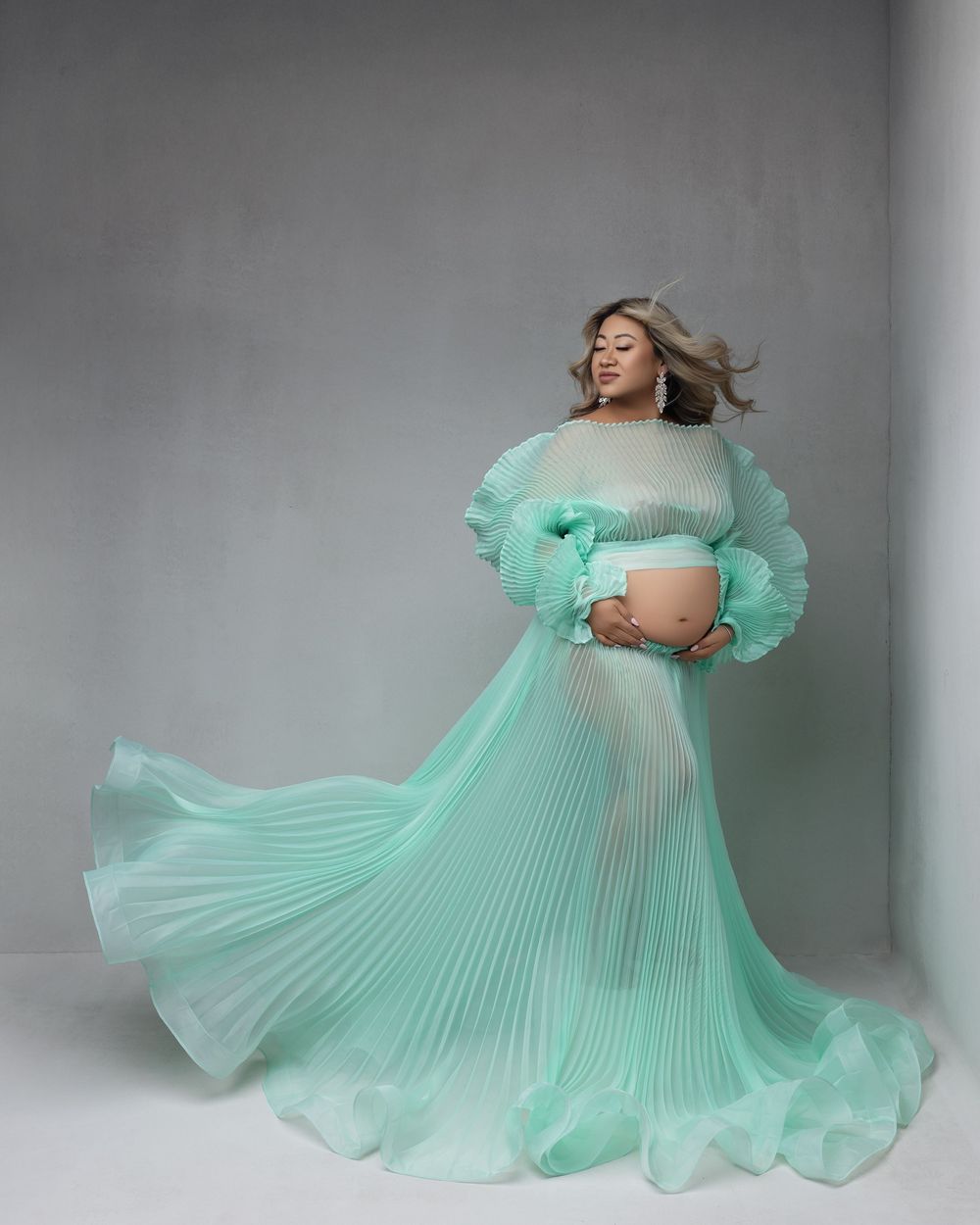 A person poses in a flowing mint green pleated maternity gown with ruffled details against a gray backdrop.