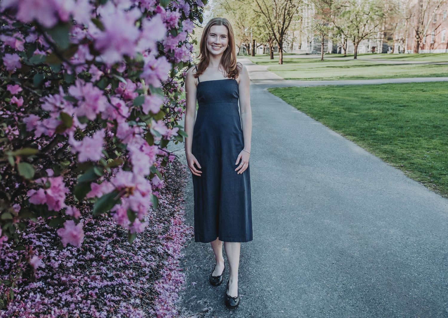A recent Bowdoin College Graduate, in a navy blue dress, stands on a path next to blooming pink cherry blossoms in a spring park setting.