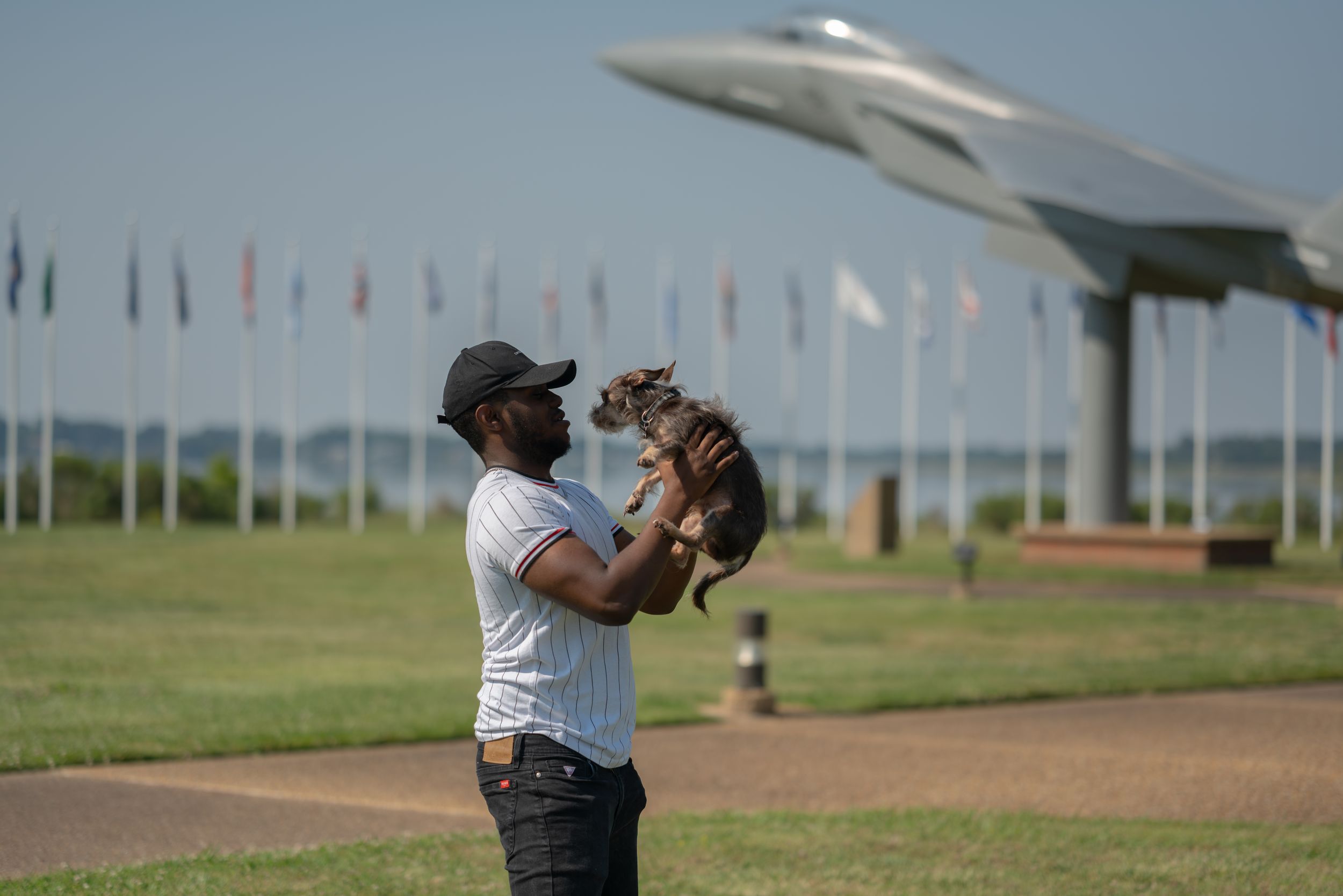 Someone holds up a small dog with a large fighter jet mounted on display in the background at an outdoor park.