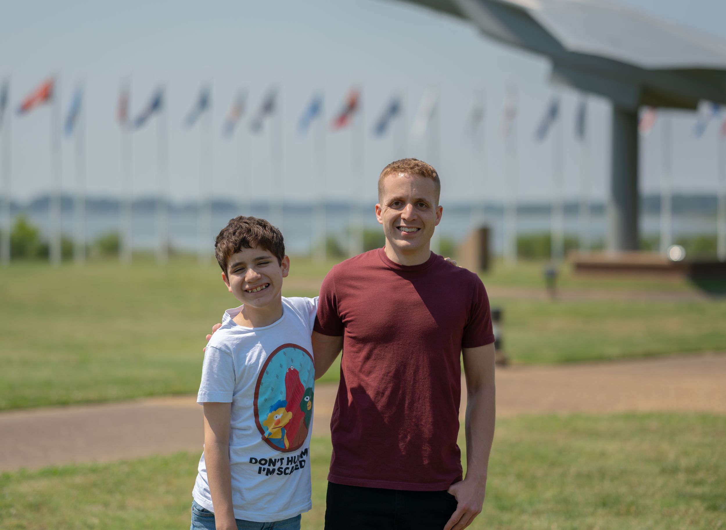 Two people stand together outdoors with colorful flags and a silver aircraft display visible in the background.