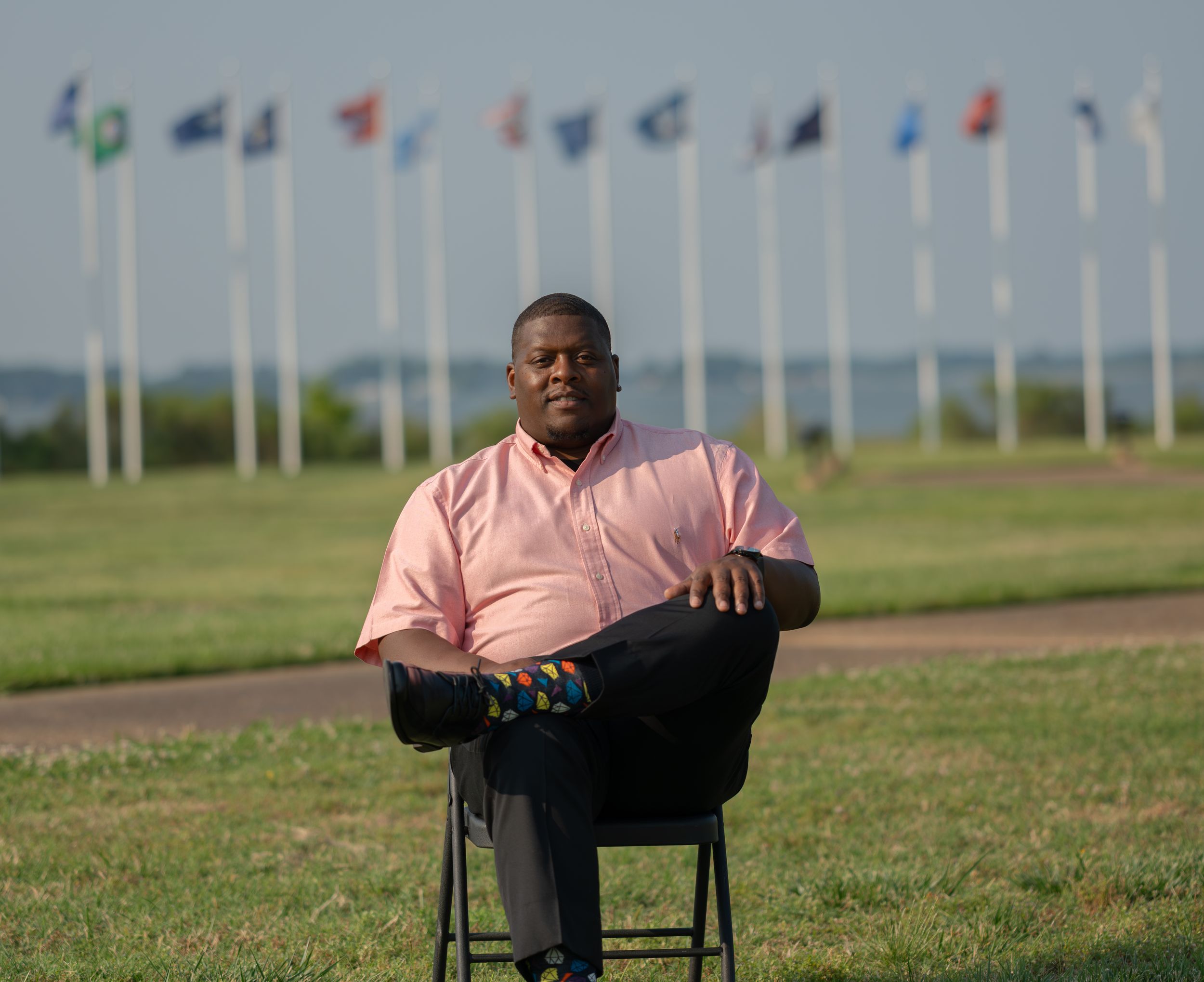 A person sits in a chair outdoors at a military memorial with rows of flags displayed behind them.