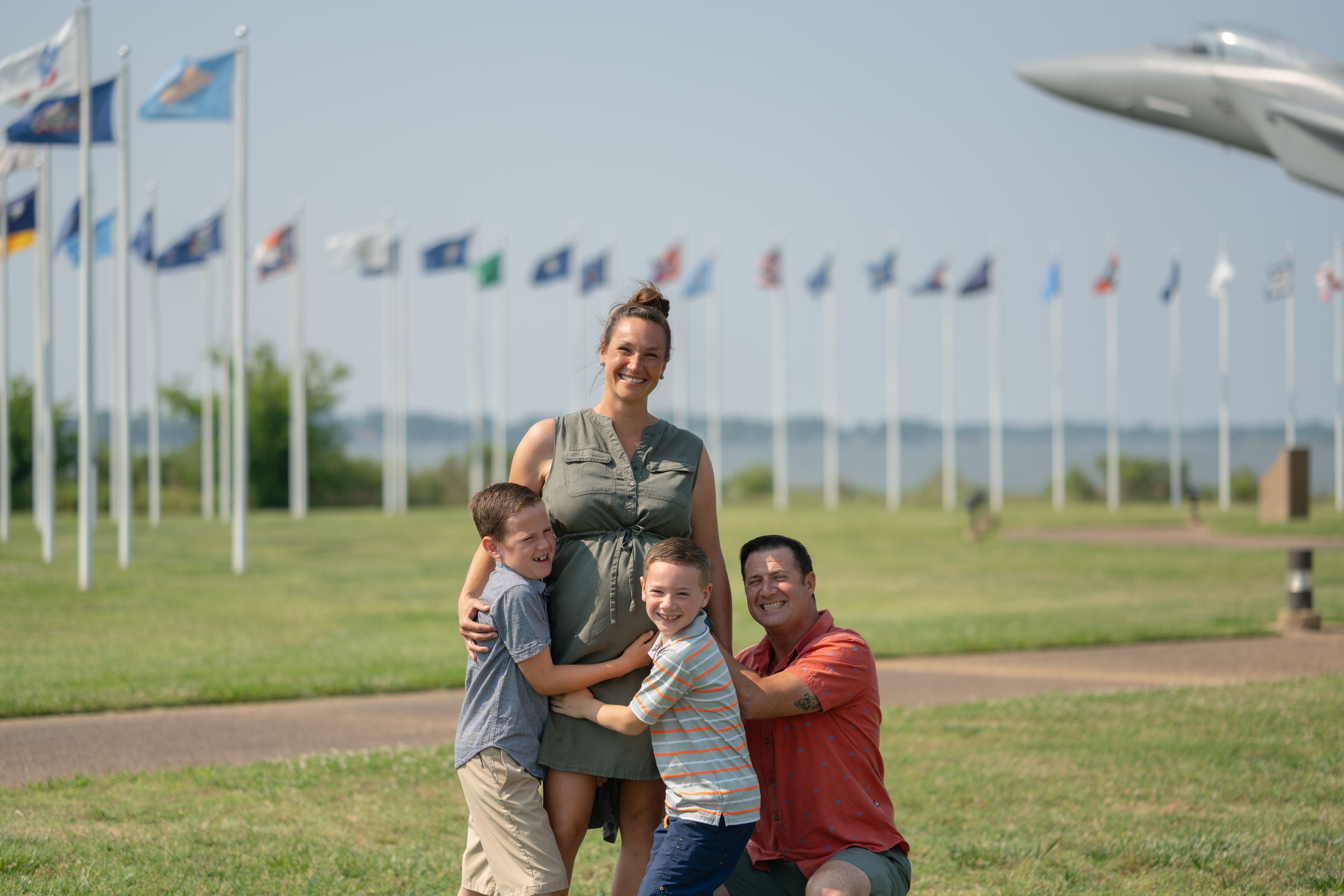 A family group embraces while posing playfully on grass with military flags and aircraft display in background.